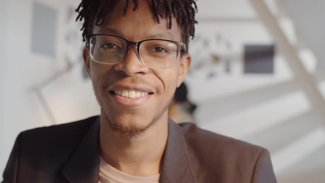 Arc chest-up shot of young handsome African American businessman in eyeglasses looking away, then turning to camera and smiling while sitting at desk in office during workday