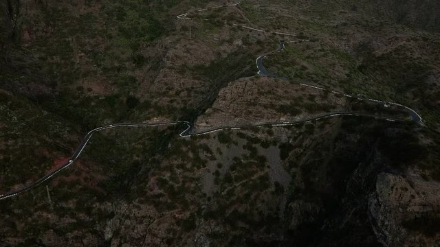 Aerial view on a serpantine road with cars. Aerial flight over serpentine road beyound mountains covered with green and brown bushes. Cars driving, slow motion