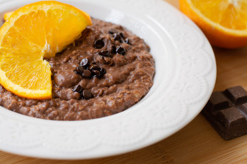 A bowl of healthy breakfast chocolate orange oatmeal porridge with chocolate chips and coconut flakes sprinkled on it. Orange and chocolate in the background. Stylish breakfast food concept.