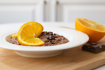 A bowl of healthy breakfast chocolate orange oatmeal porridge with chocolate chips and coconut flakes sprinkled on it. Orange and chocolate in the background. Stylish breakfast food concept.
