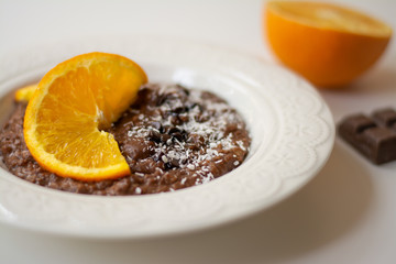 A bowl of healthy breakfast chocolate orange oatmeal porridge with chocolate chips and coconut flakes sprinkled on it. Orange and chocolate in the background. Stylish breakfast food concept.