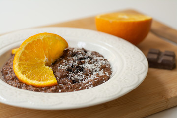 A bowl of healthy breakfast chocolate orange oatmeal porridge with chocolate chips and coconut flakes sprinkled on it. Orange and chocolate in the background. Stylish breakfast food concept.
