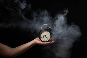 A woman holds an alarm clock in a studio full of smoke. White fog enveloped a round retro mechanical watch.