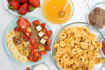 mini pancake cereal bowl with fresh strawberries banana chocolate, coconut shavings and honey in small plates. the view from the top . the trend of self-isolation