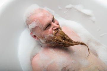 A bald man is fooling around in the foam bath. The guy makes funny hairstyles on his red beard.