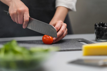 A female chef in a white uniform and a black apron in the restaurant kitchen. The cook cuts the vegetables with a large kitchen knife.