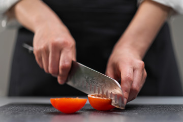 A female chef in a white uniform and a black apron in the restaurant kitchen. The cook cuts the vegetables with a large kitchen knife.