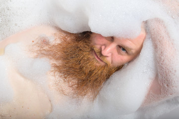 Fototapeta premium Portrait of a bald man with a long red beard takes a bath with foam. Top view on a cheerful funny guy in soapy water.