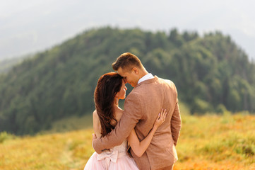 The bride and groom are standing with their backs and hugging. Sunset. Wedding photo on a background of autumn mountains. A strong wind inflates hair and dress.