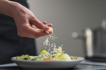 A female chef in a white uniform and a black apron in the restaurant kitchen. Cooking. The cook sprinkles grated cheese prepared food.