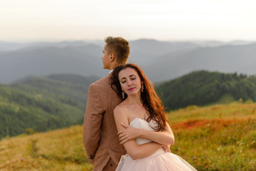 The bride and groom stand with their backs to each other. Sunset. Wedding photo on a background of autumn mountains. A strong wind inflates hair and dress. Close-up.