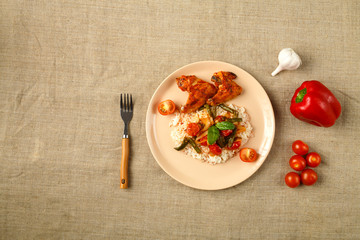 Baked chicken wings in tomato sauce with rice and vegetables on a plate on a linen tablecloth.