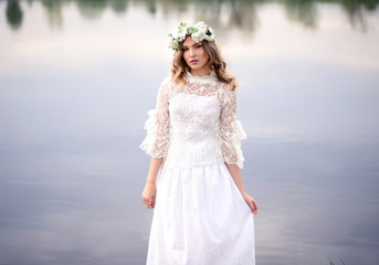 Fototapeta na wymiar A beautiful girl in a white dress with a wreath of flowers stands near the river. Young girl on the background of a blue lake