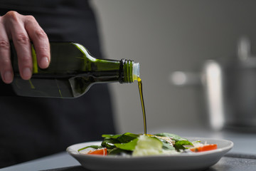A female chef in a white uniform and a black apron in the restaurant kitchen. Cooking. The cook pours olive oil from a green bottle.