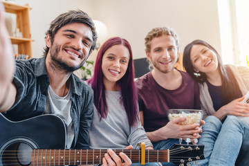 Smiling friends in casual on couch at home with guitar making selfie
