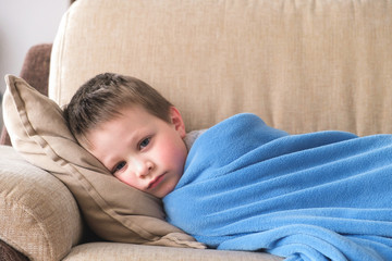 A sick or sad little boy of preschool age lies on a sofa wrapped in a blue plaid. Children's...