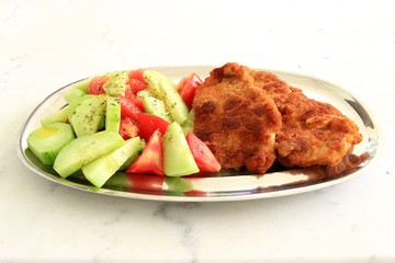 Fried breaded chicken wiener schnitzel with tomato and cucumber fresh salad in a metal dish on a...
