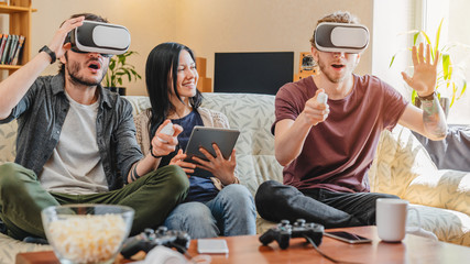 Amazed friends having fun with VR googles at home while sitting on sofa