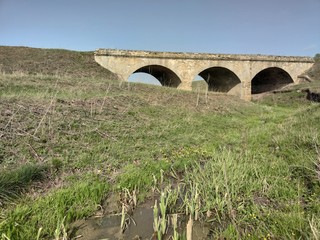 Old soviet bridge. Little gray river. Green grass. Blue sky.