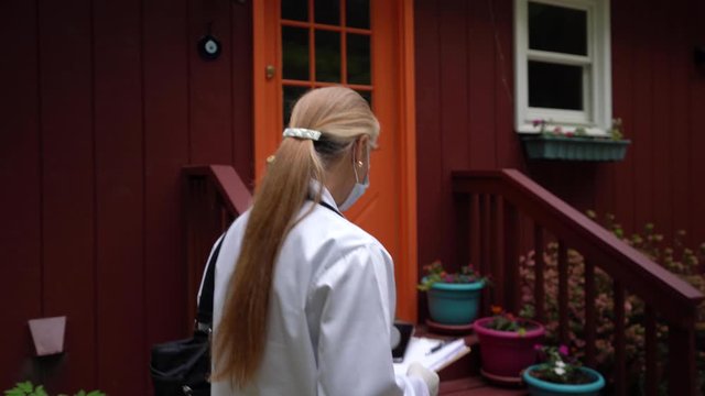 Camera follows behind a nurse or doctor going to a house for home visit wearing face mask and gloves, knocks and enters door.