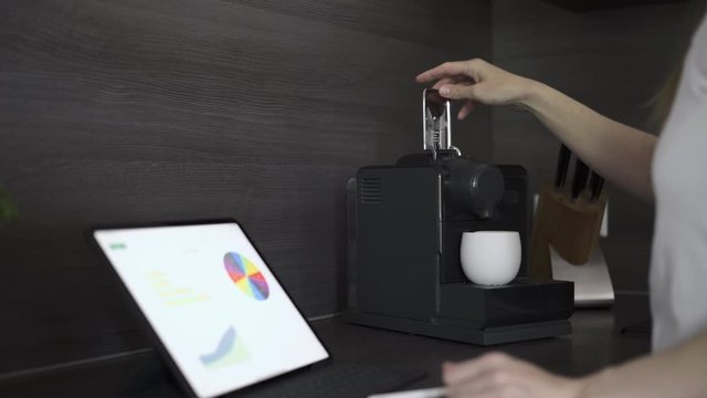 Frau arbeitet im home office zu Hause am Tablet, telefoniert mit Bluetooth Headset und macht sich dabei einen Kaffee 