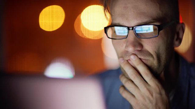 Closeup of focused young man in eyeglasses facing difficulties while working on laptop at night, pc screen reflecting in his eyewear