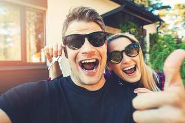 young happy couple with keys in hand standing outside of their new home and take a selfie