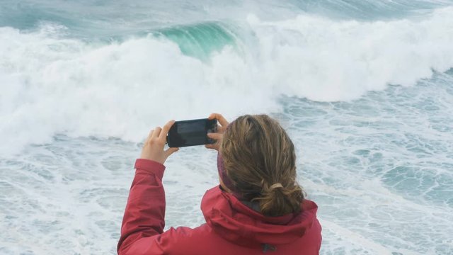 girl in pink jacket takes photos on smartphone of giant ocean waves slow motion, sea in a storm