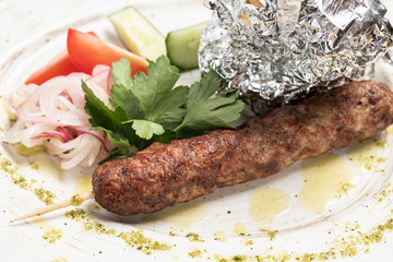 Lula kebab on a skewer with vegetables tomatoes and cucumber slices on a white plate