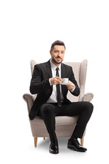 Young professional man in suit and tie sitting in an armchair abd holding a cup of espresso coffee