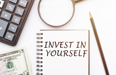 Top view of calculator, magnifying glass, dollars and note written with 'Invest In Yourself' on white background.