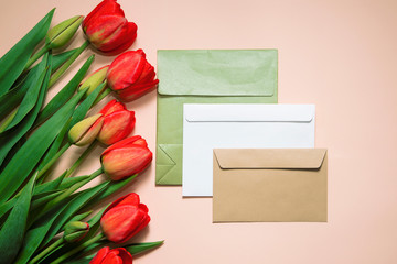 Three envelopes of different colours and tulips on a beige background. Top view, flat lay.