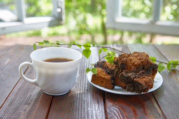 Chaga mushroom and a cup of tea stand on a wooden surface next to an open window on a summer sunny day.