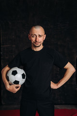 A male sports teacher stands against a dark background, holding a soccer ball