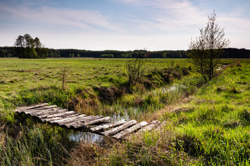 Wooden pedestrian bridge over canal ditch with water. Countryside nature serenity meadow landscape view in Zdzarka, Poland, Europe.