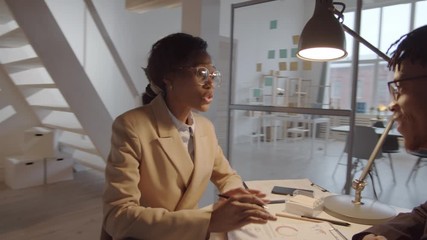 Full 360 tracking shot of African American female manager discussing business with young male colleague while sitting together at desk in office