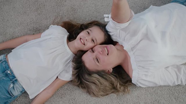 A beautiful mother and daughter lie on the floor inside the house and look up, smiling and having fun chatting. Top view