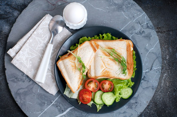 Hot sandwiches with cream cheese, cucumber, ham, tomatoes and peas sprouts on black plate. Breakfast with egg mist on white stand. Top view