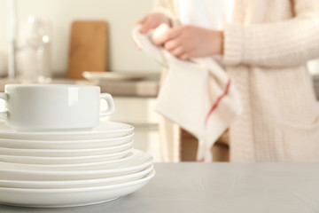 Woman wiping plate with towel in kitchen, focus on stack of clean dishes
