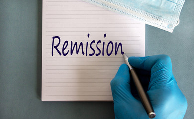 Male doctors hand in blue glove writes the word 'remission' on a white note. Medicine mask....