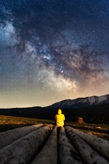 A man in yellow jacket sitting on a trees staring at a starry sky with a Milky Way and a Tatra...