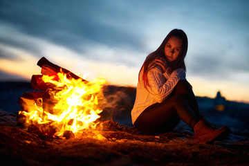 The girl traveler in the evening warms himself by the fire in the mountains near the camp, the girl is lit by the light of fire. Sunset in the background