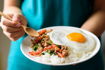 Thai food, stir-fried holy basil with minced pork and fried egg (Pad Kaprao Moo Kai Dao) on plate holding by hand and eating
