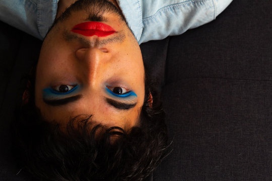 Non-binary bearded young Hispanic man on a gray background, with flamboyant makeup posing, red lips and blue eye shadows