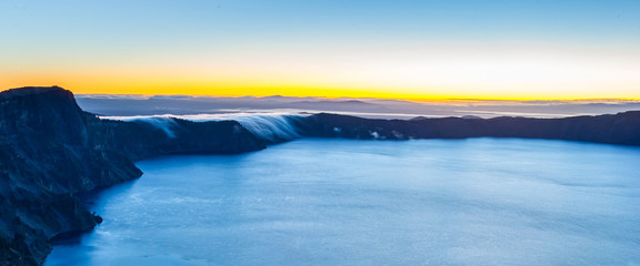 The sun rises on the horizon as low morning clouds flow like water over the rim and down the side of the caldera to the large lake in the crater below.