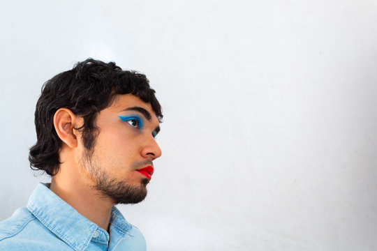 Non-binary Bearded Young Hispanic Man On A White Background, With Flamboyant Makeup Posing, Red Lips And Blue Eye Shadows