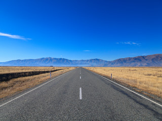 Straight empty Country Road leading to Mountains lining the horizon on sunny day, Otago region, South Island, New Zealand.