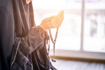 Close up beautiful asian muslim woman palms open to light praying to allah with prayer beads in the hands of the palm, wearing black hijab robe, in a private prayer room with bright light warm tone