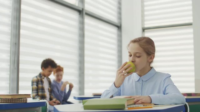 Medium shot of blond-haired Caucasian girl sitting at desk in school, reading textbook and eating green apple while two her classmates talking on background