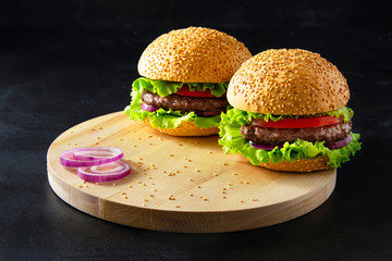 Two homemade burgers and onion rings on little wooden cutting board over dark background.